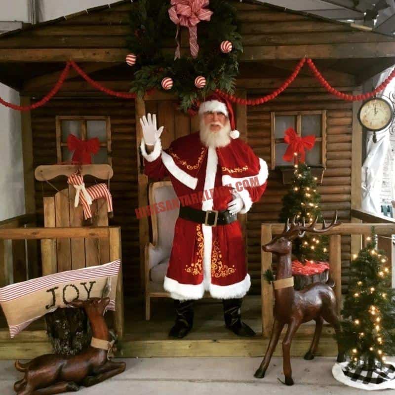 Real bearded Santa waving from a log cabin porch with reindeer figures and a wreath