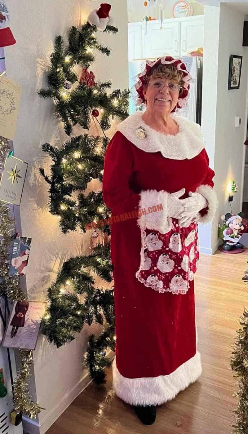 Mrs. Claus for hire in a red velvet dress and bonnet beside a lit Christmas tree