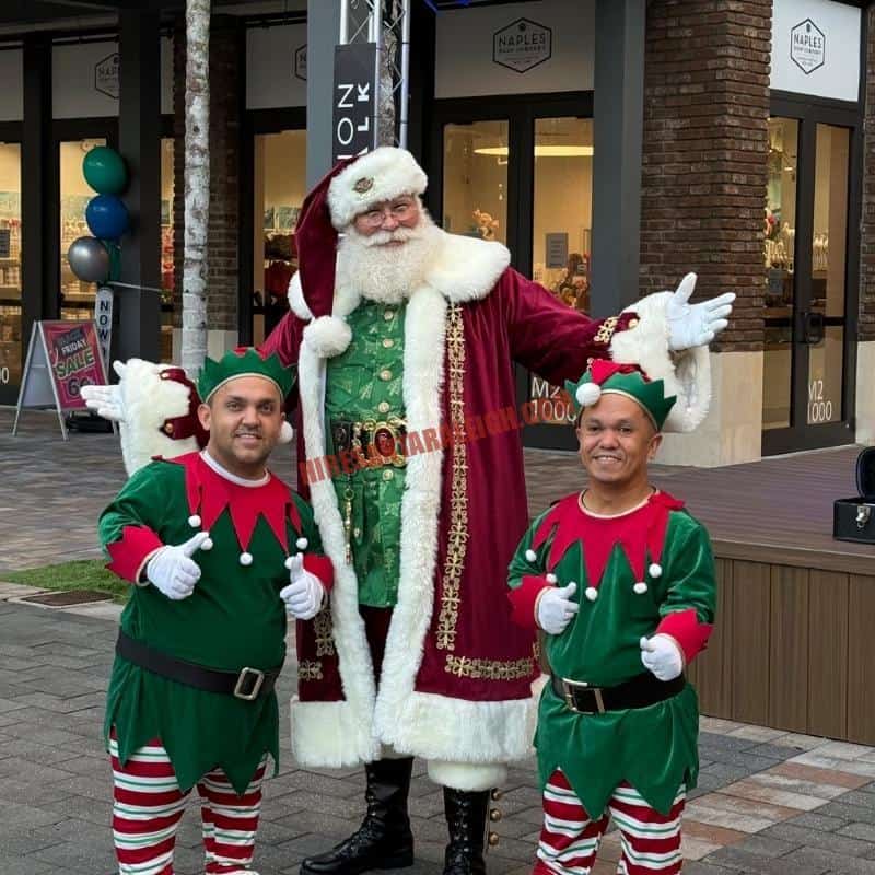 Santa Claus impersonator in a red and green coat posing outdoors with two elves